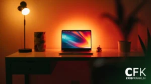 Um laptop repousa sobre uma mesa, iluminado por uma luminária aconchegante. Há uma xícara estampada, uma pequena estatueta e uma planta, com as iniciais "CFK" e "Cris Franklin" no canto inferior direito.