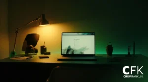 Uma mesa mal iluminada com um laptop, luminária, telefone e vários itens; a tela exibe um site e a sala tem iluminação verde e amarela. Logotipo "CFK CRISFRANKLIN" no canto inferior direito.