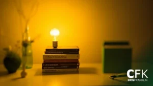 Uma lâmpada acesa sobre uma pilha de livros sobre uma mesa, com uma planta desfocada e uma caixa ao fundo. A imagem tem uma iluminação amarela suave.
