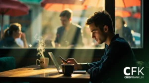 Um homem senta-se à mesa de um café usando um laptop e um smartphone, com a luz do sol entrando e uma xícara fumegante por perto; outras pessoas são visíveis ao fundo.