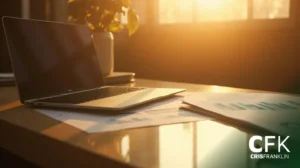 Um laptop e documentos descansam em uma mesa de madeira sob a luz do sol, com uma planta e uma janela ao fundo. O logotipo da CFK (Cris Franklin) está visível no canto inferior direito.