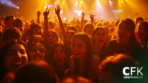Uma grande multidão de jovens sorri e aplaude em um show ou evento interno bem iluminado, com luzes de palco laranja ao fundo. As letras "CFK Cris Franklin" estão visíveis no canto inferior direito.