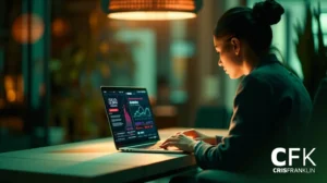 Uma mulher senta-se em uma mesa em uma sala pouco iluminada, trabalhando em um laptop que exibe dados financeiros e gráficos de ações. A imagem apresenta a marca d'água "CFK CRISFRANKLIN" no canto.