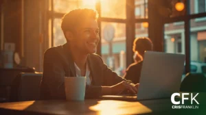 Uma pessoa senta-se à mesa em um café iluminado pelo sol, sorrindo enquanto usa um laptop. Uma xícara de café branca está sobre a mesa. As letras "CFK" aparecem no canto inferior direito.