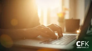 Uma pessoa digita em um teclado de laptop em uma sala iluminada pelo sol, com uma xícara embaçada e flores ao fundo. O logotipo da CRISFRANKLIN aparece no canto inferior direito.