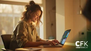 Uma mulher sentada em uma mesa trabalhando em um laptop em uma sala iluminada pelo sol, com uma caneca de café por perto. O logotipo "CFK CRISFRANKLIN" está visível no canto inferior direito.