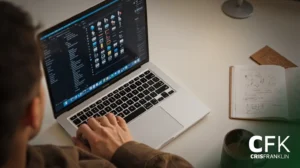 Pessoa usando um laptop com uma interface de tema sombrio, caderno aberto, caneta e caneca de café em uma mesa. A imagem inclui uma marca d'água CFK CRISFRANKLIN.