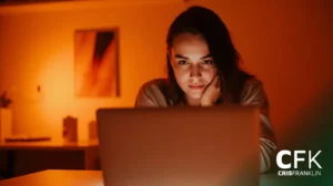 Uma pessoa está sentada em uma mesa em uma sala mal iluminada, olhando atentamente para a tela de um laptop. As letras "CFK" e "Cris Franklin" aparecem no canto inferior direito.