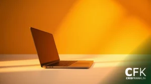 Um laptop fino está aberto em uma superfície branca contra um fundo amarelo, com sombras projetadas pela luz. "CFK CRISFRANKLIN" está visível no canto inferior direito.