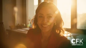 Uma mulher vestindo um blazer vermelho sorri enquanto está sentada em uma sala iluminada pelo sol com grandes janelas. O logotipo "CFK CRISFRANKLIN" está visível no canto inferior direito.