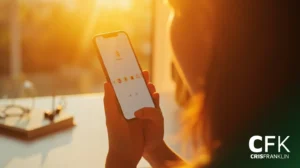 Uma pessoa segura um smartphone que exibe uma tela de login com ícones de emoji, sentada em uma mesa iluminada pelo sol, com óculos visíveis ao fundo.