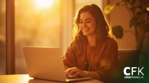 Uma mulher sentada em uma mesa usando um laptop, sorrindo, com a luz quente do sol passando por uma janela; o logotipo "CFK CRISFRANKLIN" aparece no canto inferior direito.