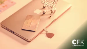 Um cartão de crédito e um pequeno carrinho de compras de arame repousam sobre um laptop fechado com um chaveiro próximo. A imagem tem a marca "CFK CRISFRANKLIN" no canto inferior direito.