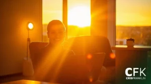 Uma pessoa sentada dentro de casa com um laptop sorri para a câmera enquanto a luz do sol atravessa uma janela ao pôr do sol. O logotipo "CFK CRISFRANKLIN" pode ser visto no canto inferior direito.